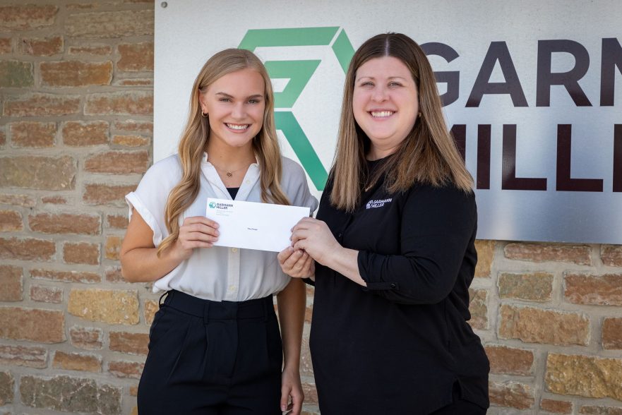 Garmann Miller team member Juliana hands scholarship check to Macy Prenger in front of Garmann Miller office sign